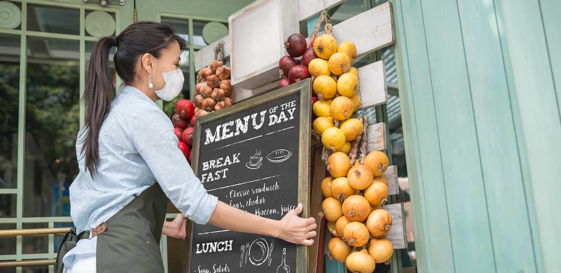 A business owner wearing a mask and placing a menu outside of their cafe.