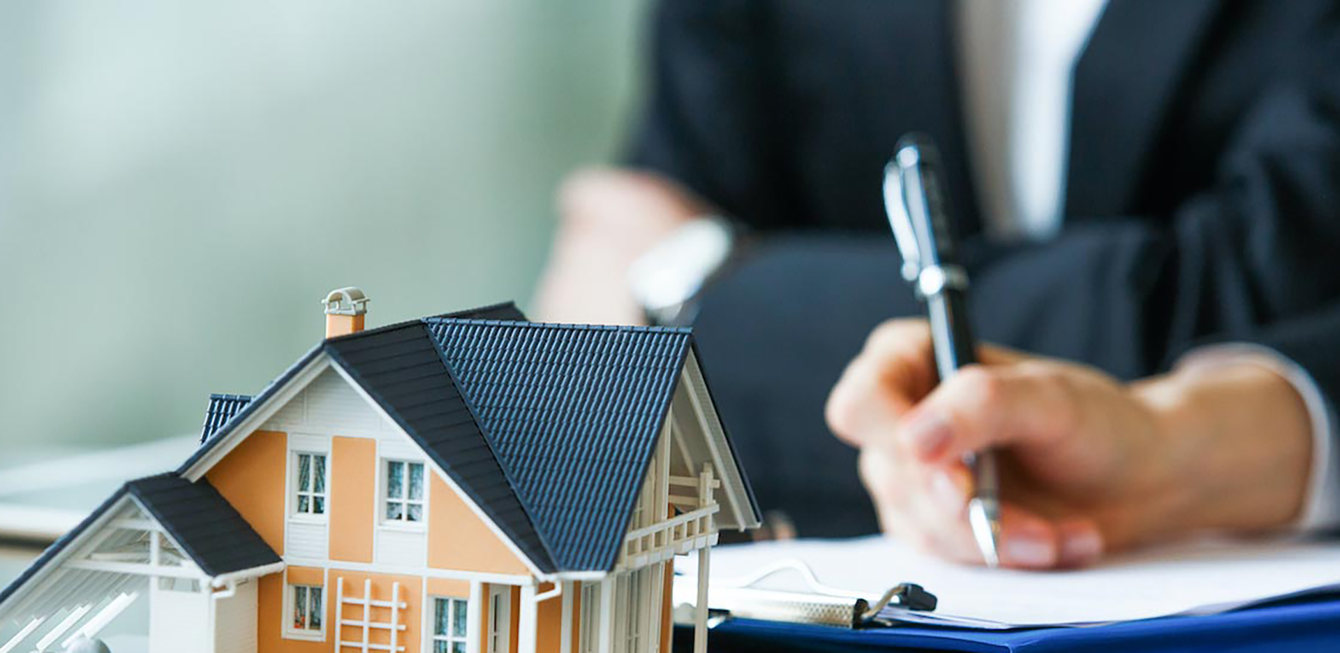 A small model of a home in front of someone signing documents on a clipboard.