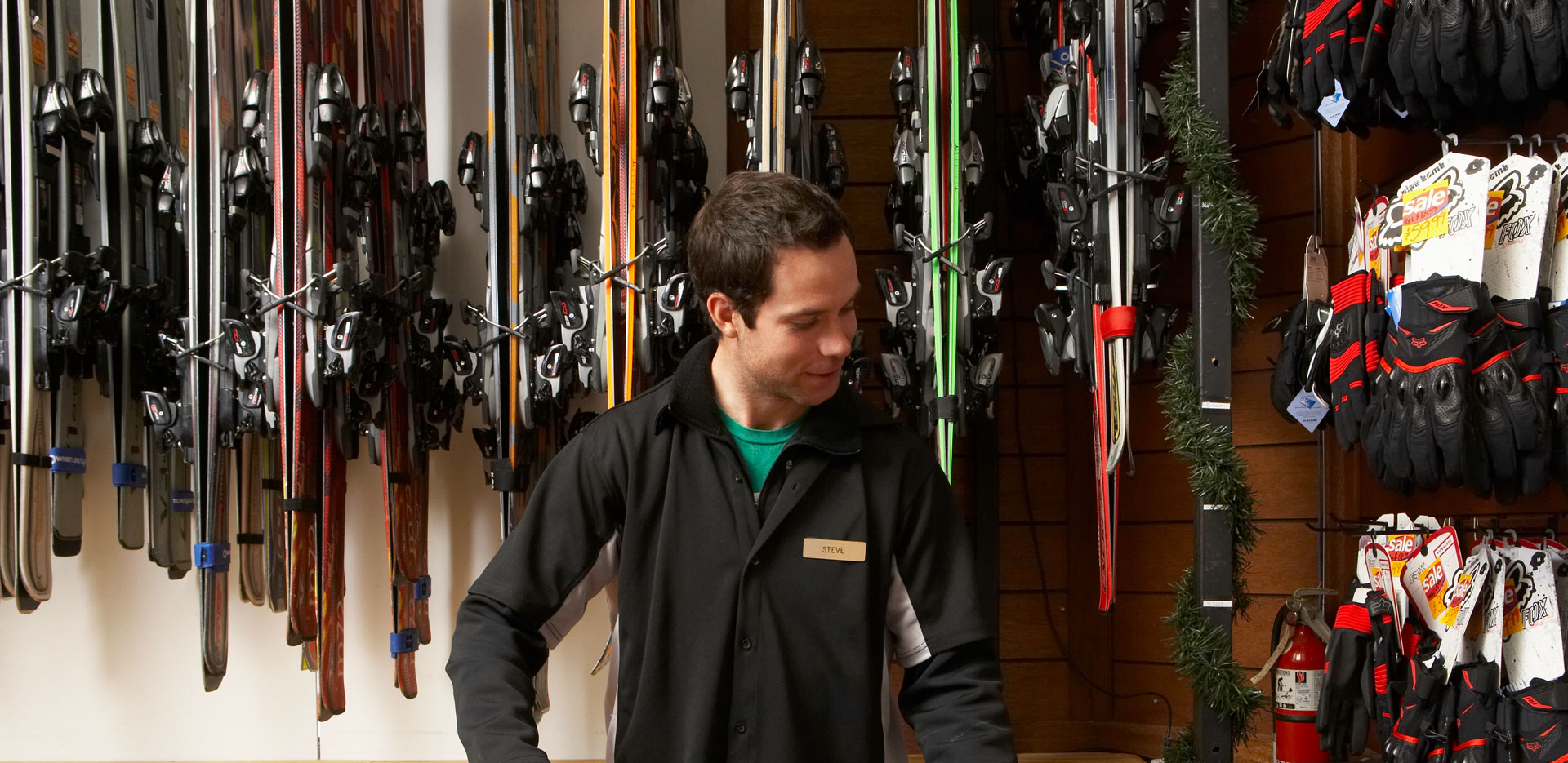 A man wearing a nametag standing in front of a wall of skis.