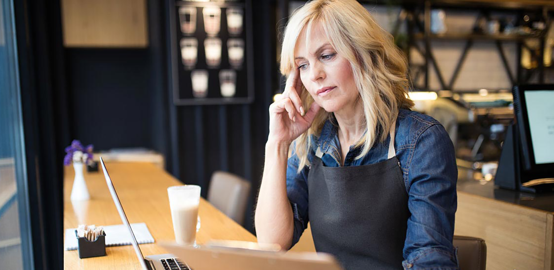 A cafe owner wearing an apron clutching her head while working on her laptop.