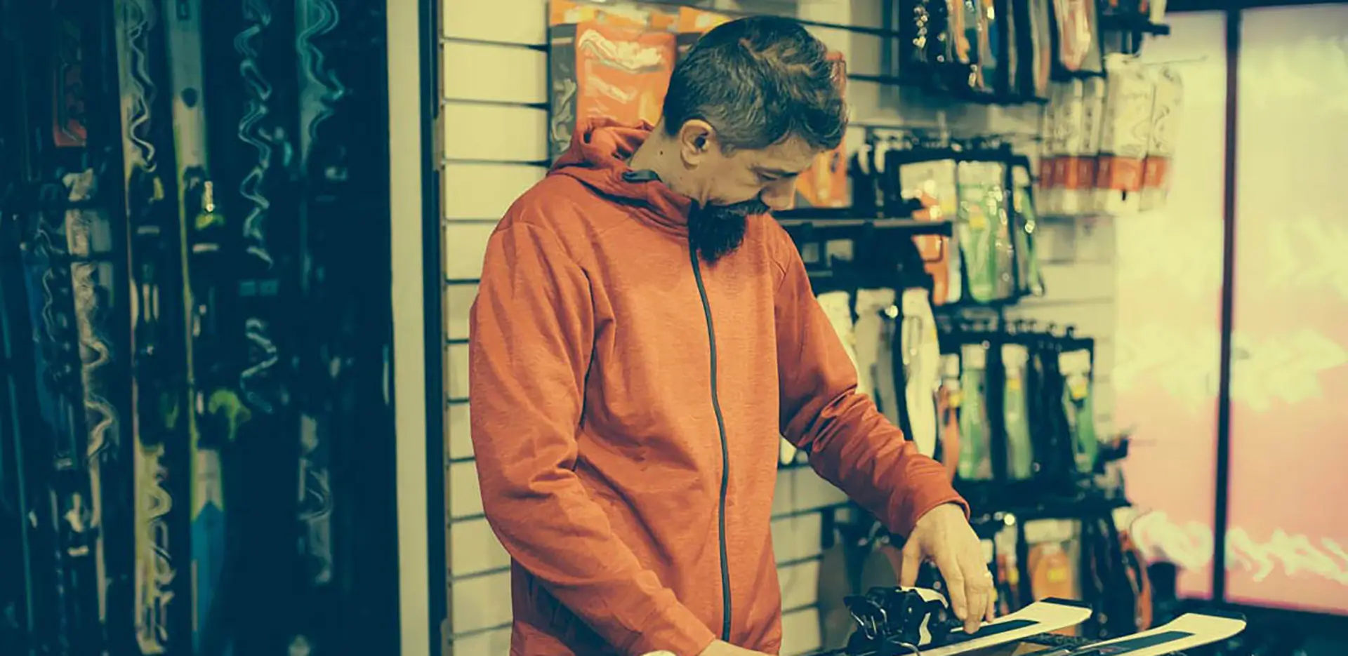 A man in an orange sweater repairing a pair of skis in a ski shop.