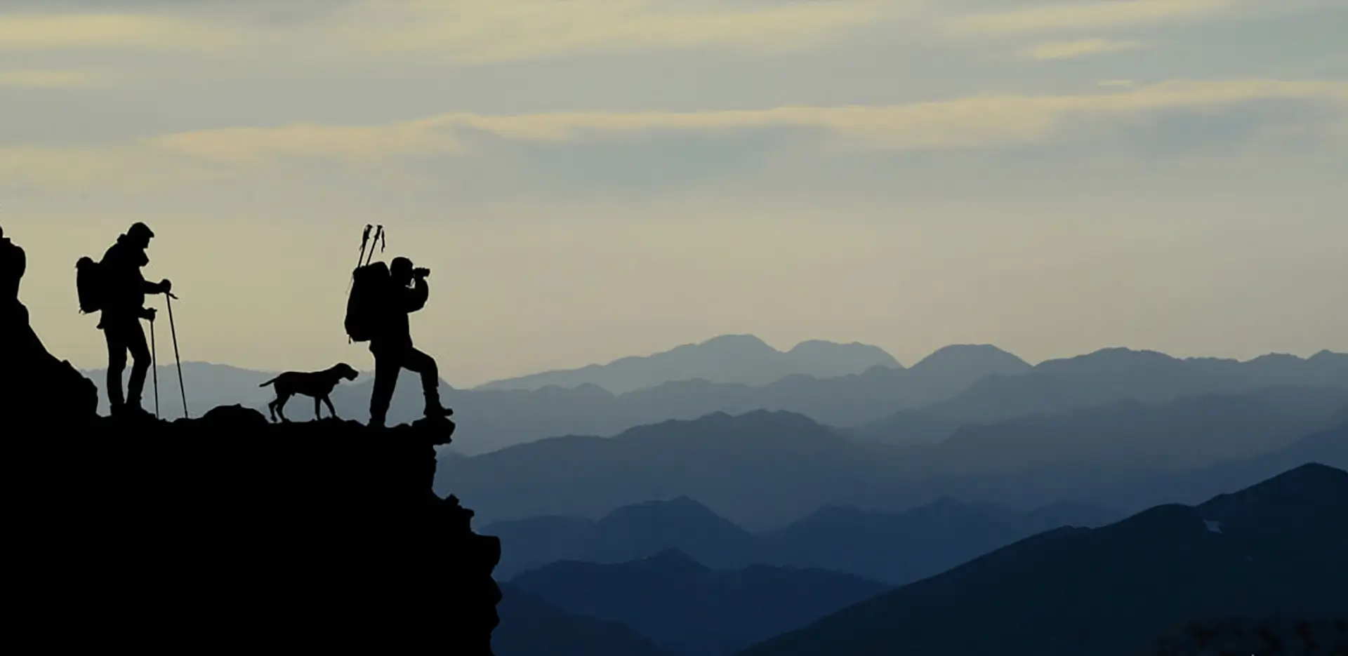 Two hikers and a dog in silhouette standing at the top of a cliff.