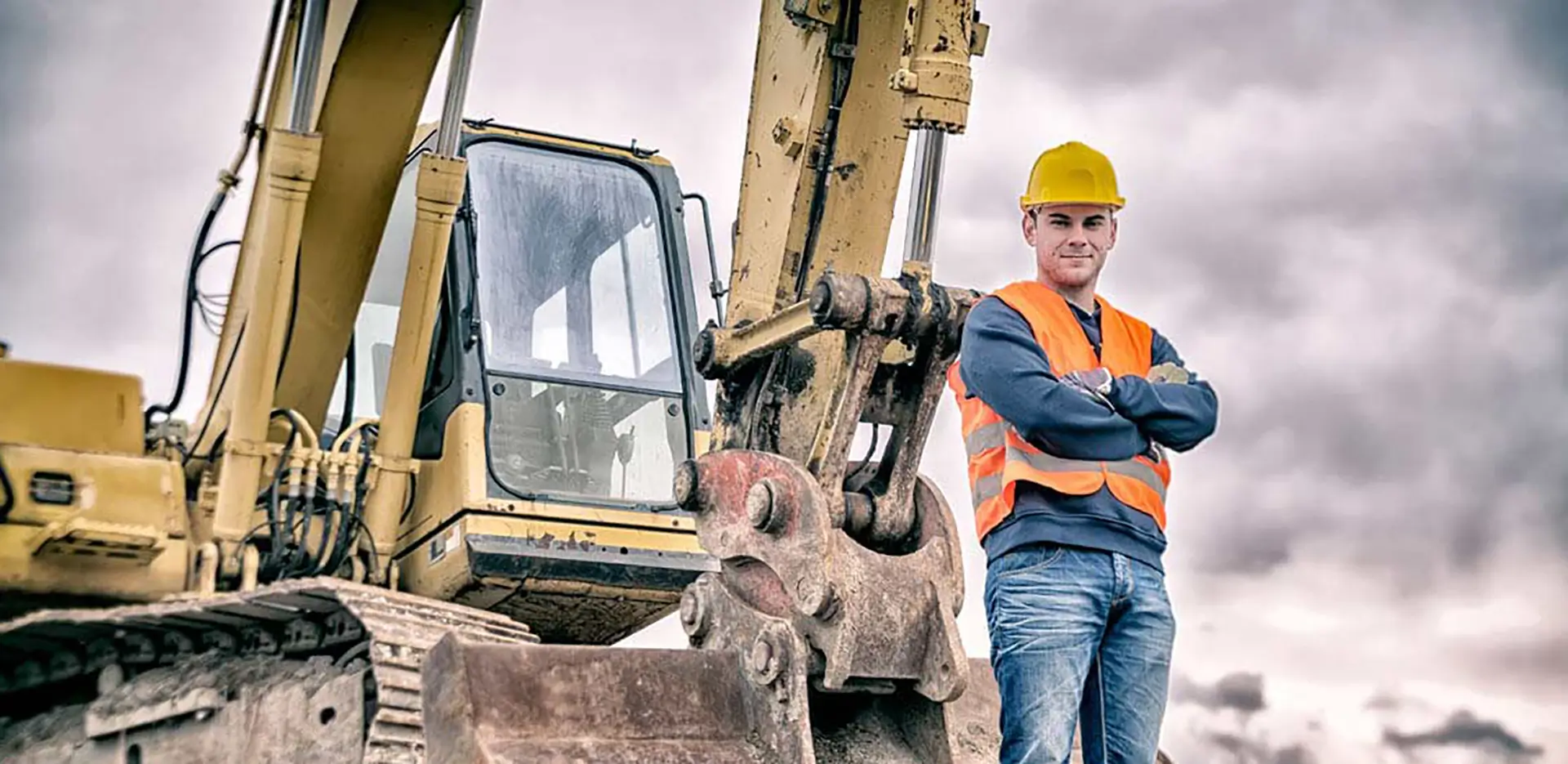 A construction worker standing with his arms folded in front of an excavator.