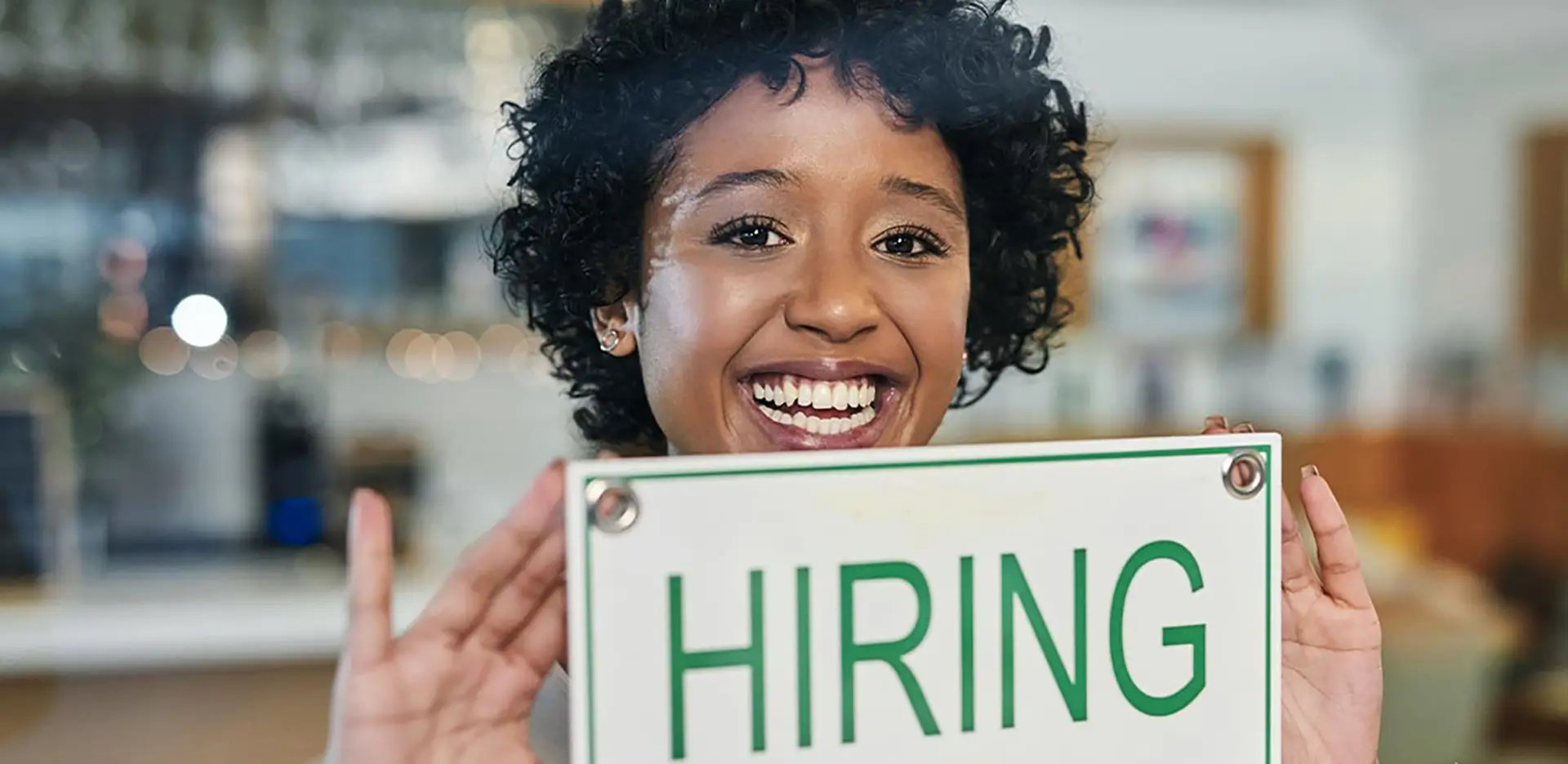 A smiling woman hanging a 'hiring' sign on the door of her business.