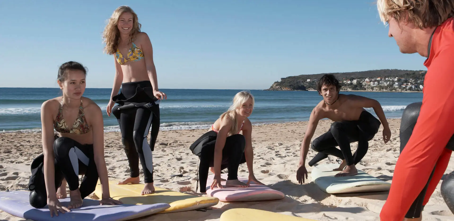 A group of teens practicing standing on a surfboard on the beach.
