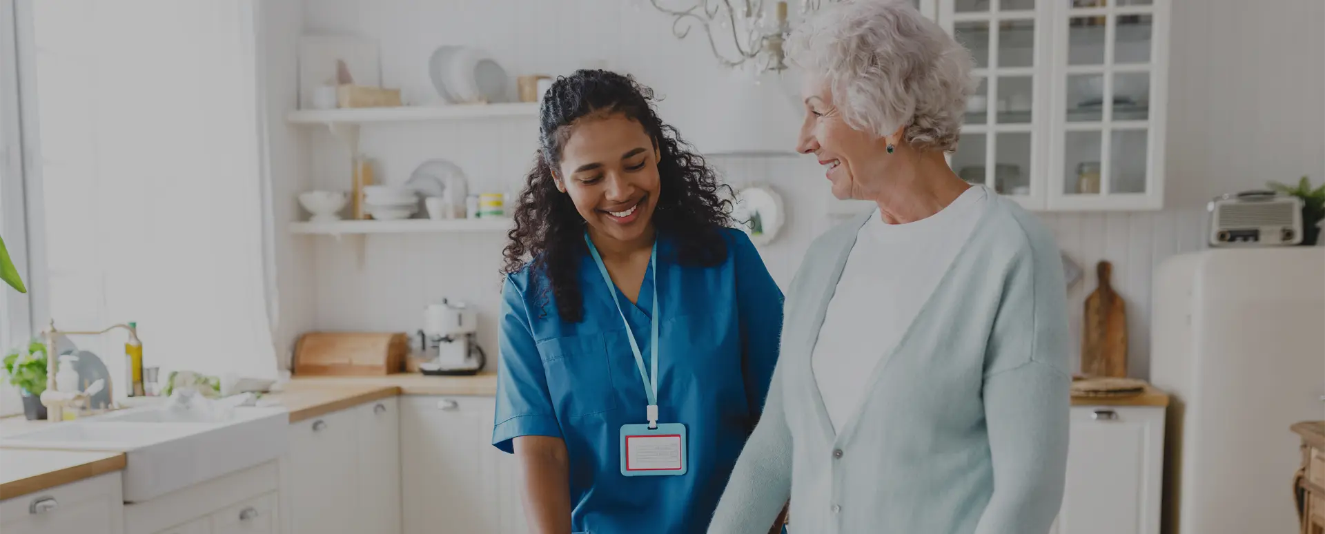 A smiling nurse helps and elderly woman with a walker in a house kitchen.