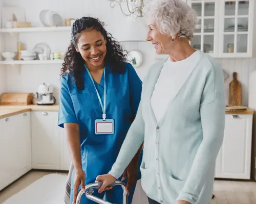 A smiling nurse helps and elderly woman with a walker in a house kitchen.