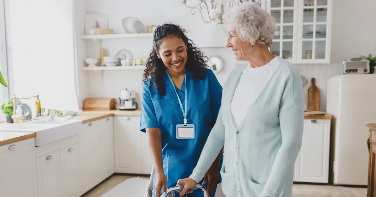 A smiling nurse helps and elderly woman with a walker in a house kitchen.