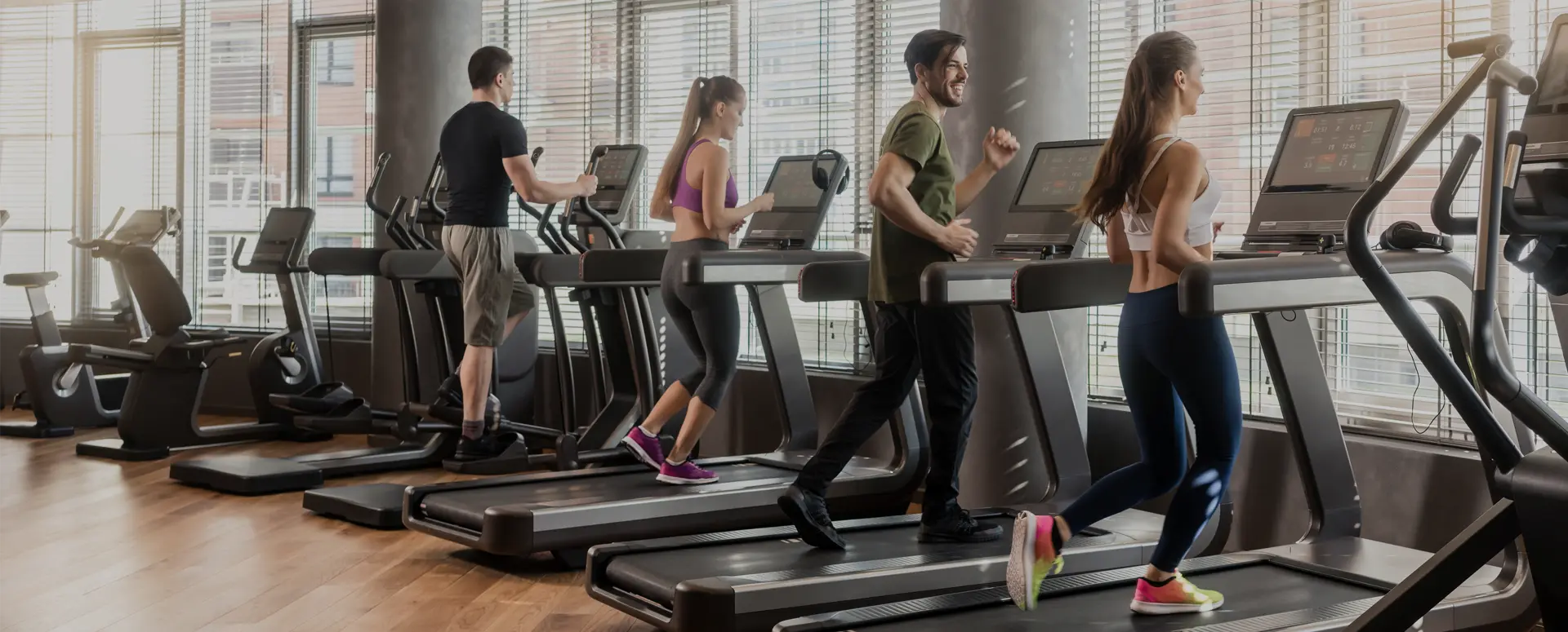 People running on treadmills at a gym with large windows along the wall.
