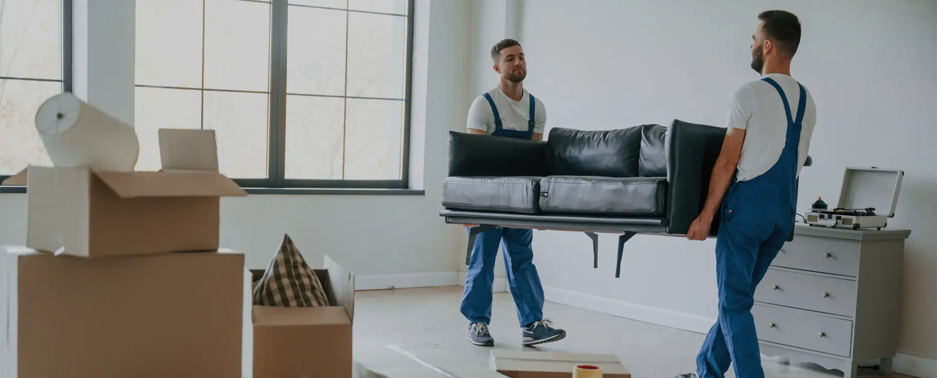 Two men wearing overalls carry a couch through a room filled with boxes.