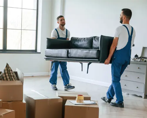 Two men wearing overalls carry a couch through a room filled with boxes.