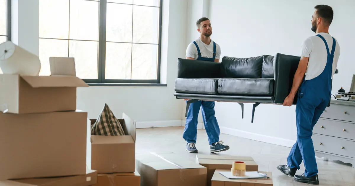 Two men wearing overalls carry a couch through a room filled with boxes.