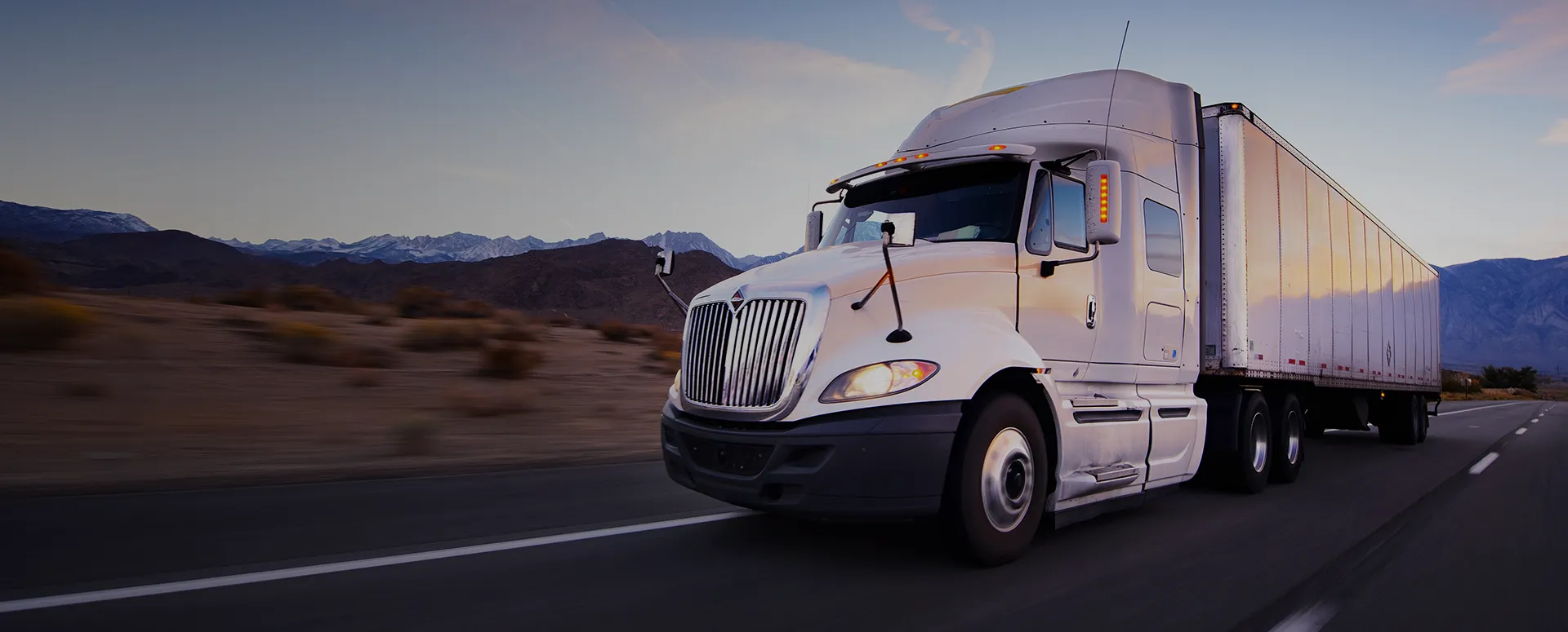 A white semi-truck driving along a desert road with mountains in the distance.