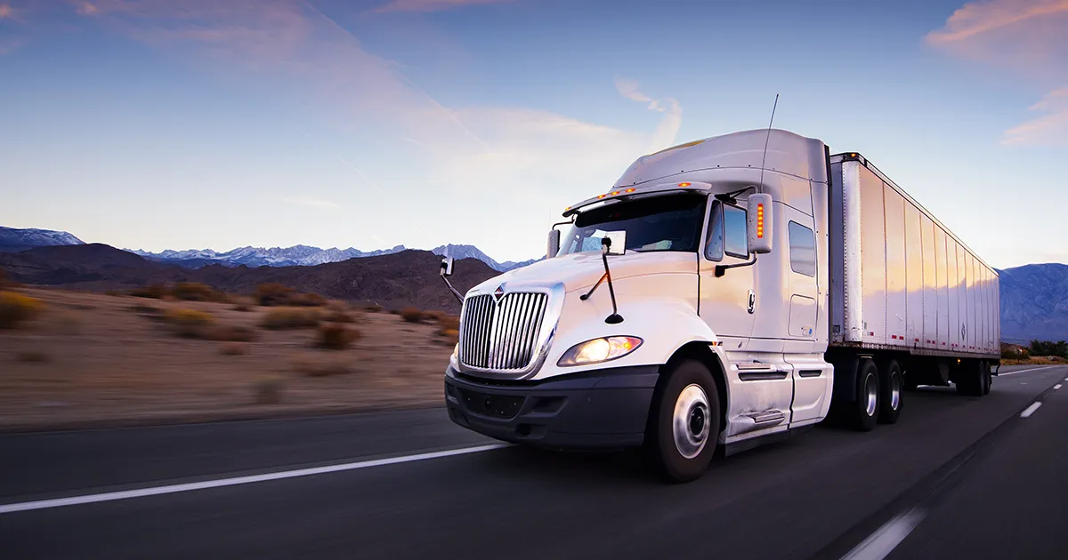 A white semi-truck driving along a desert road with mountains in the distance.