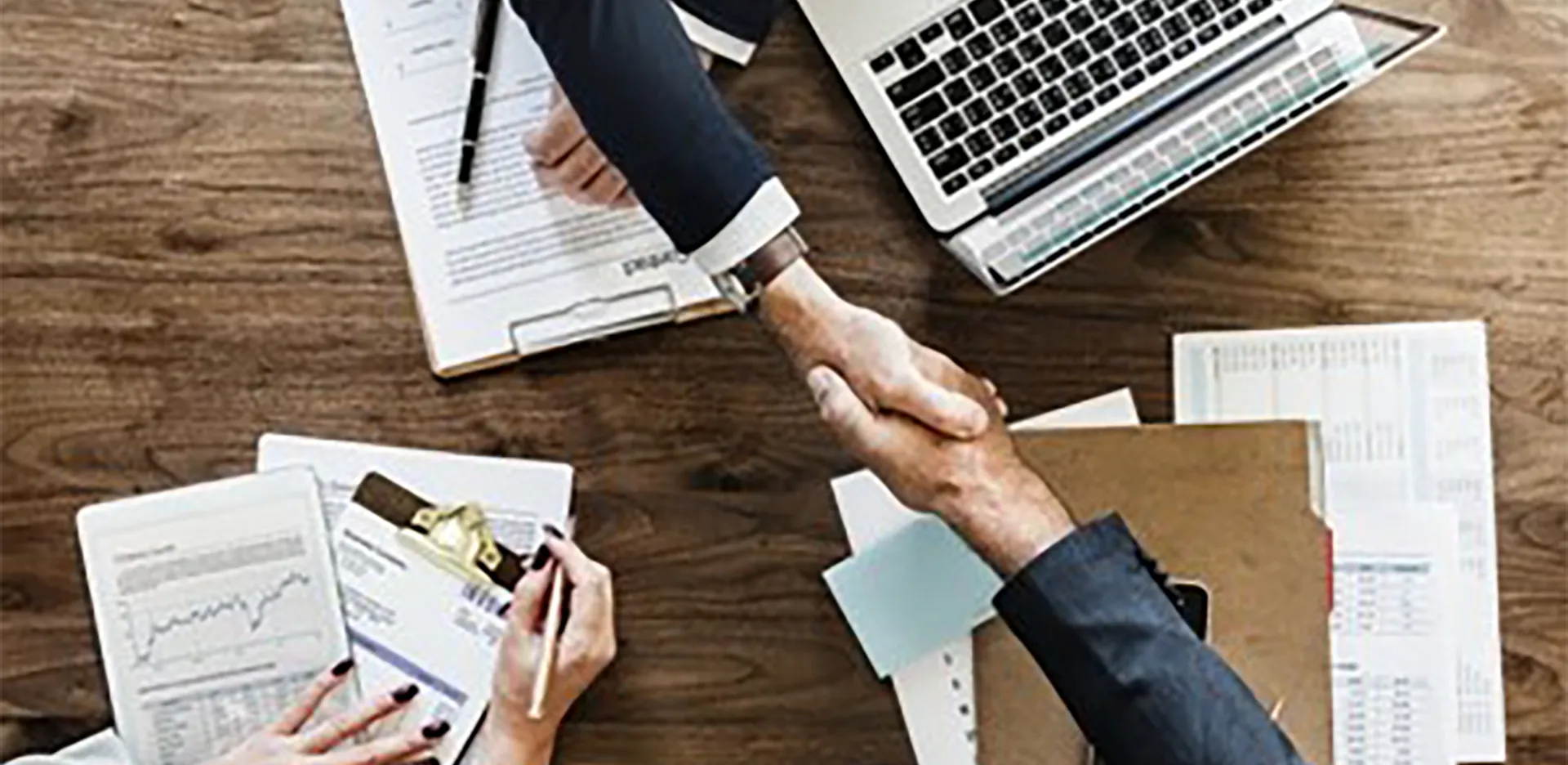 Two hands shaking against a background of documents and a laptop computer.