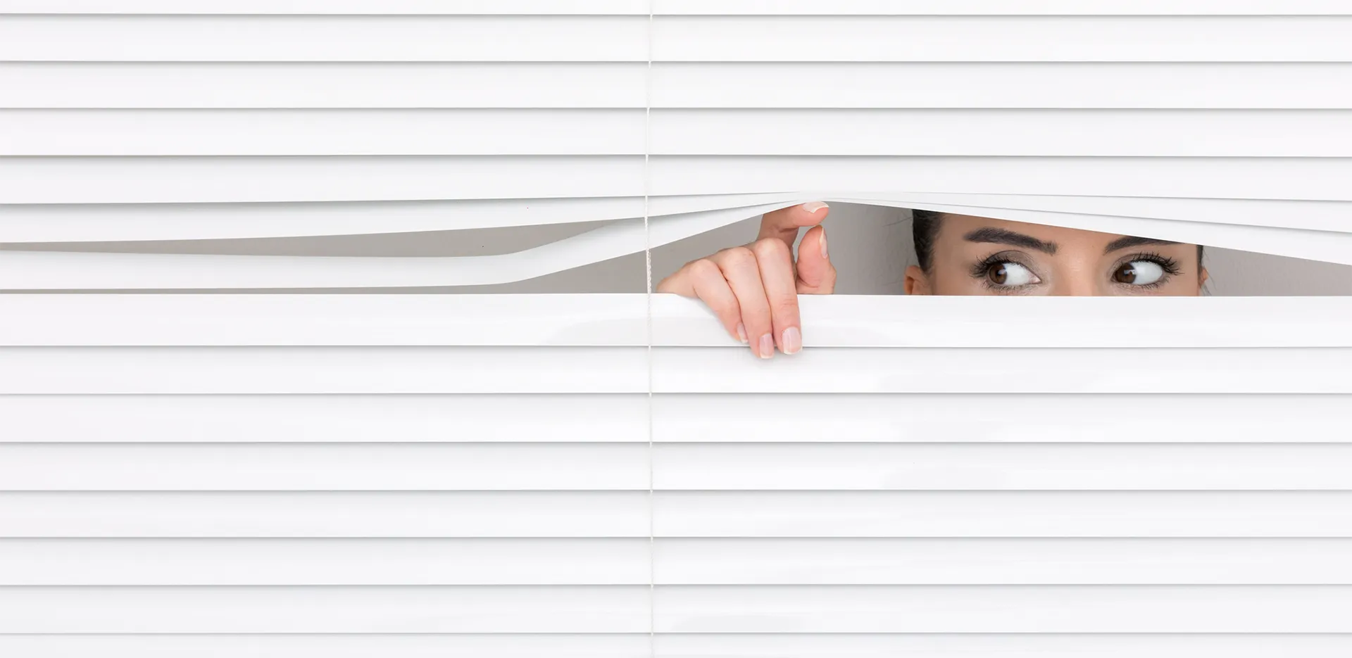 A woman peeking through the slats in a set of blinds.