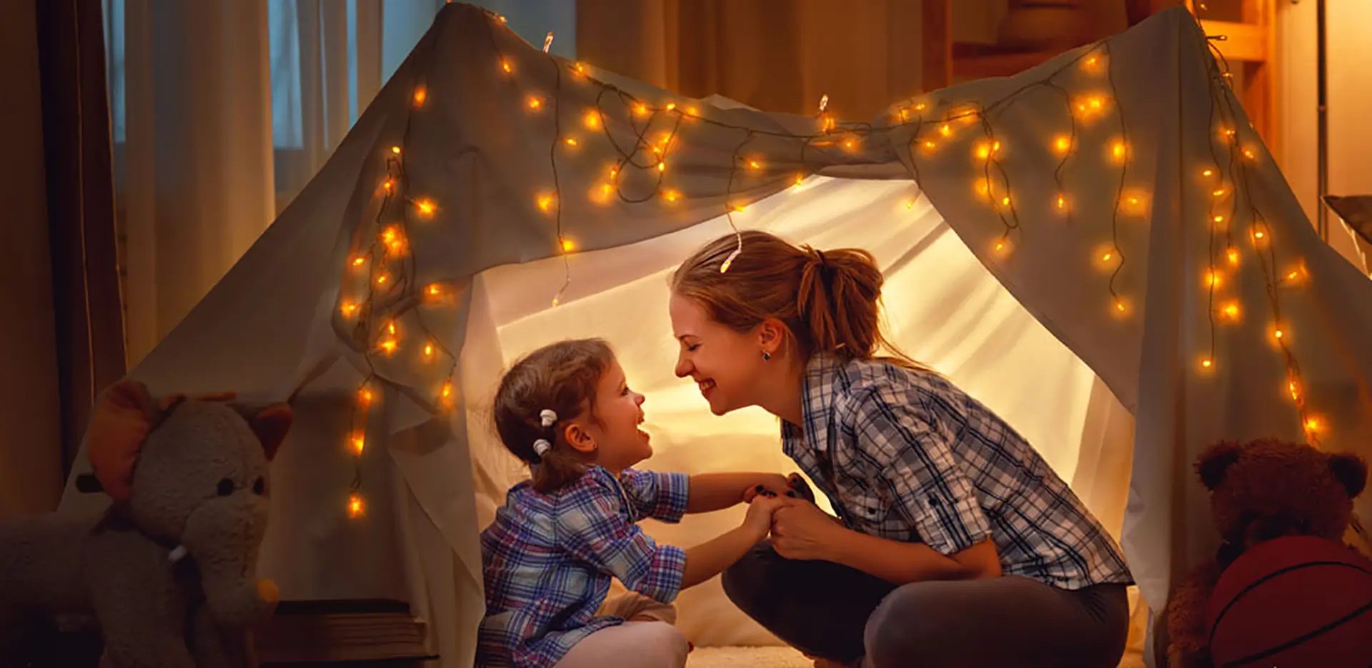 A mom and her daughter smiling and laughing in a blanket fort.