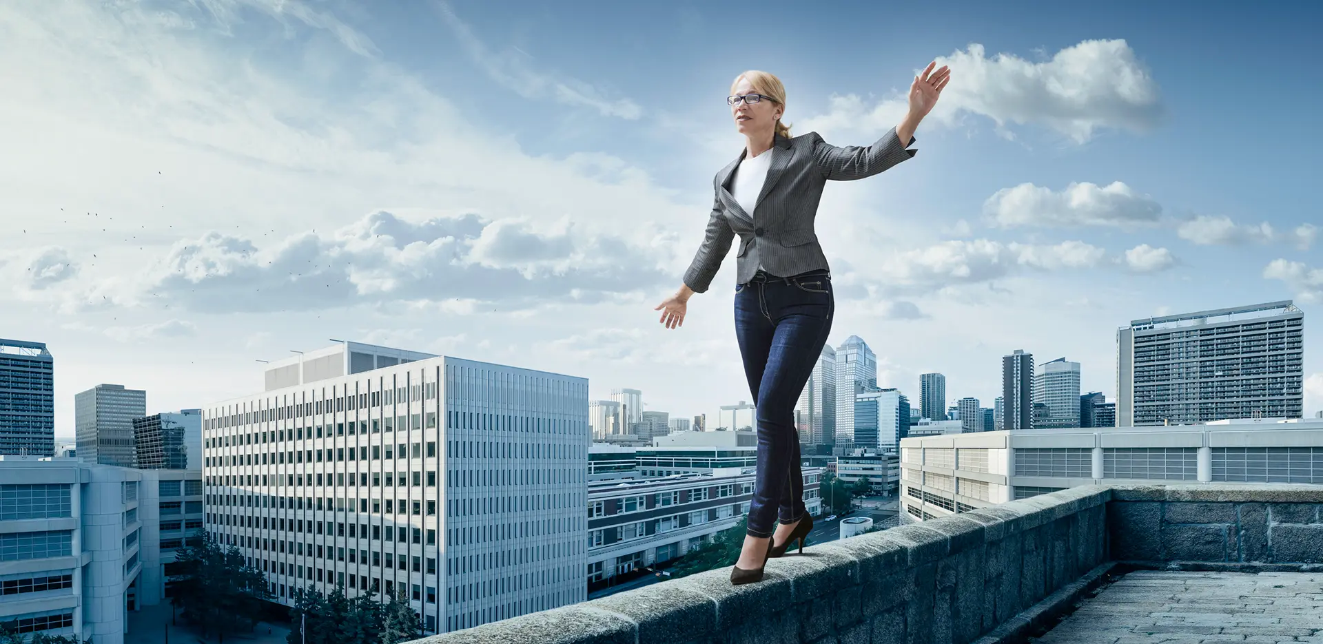 A woman balancing on the ledge of a tall building with a city behind her.
