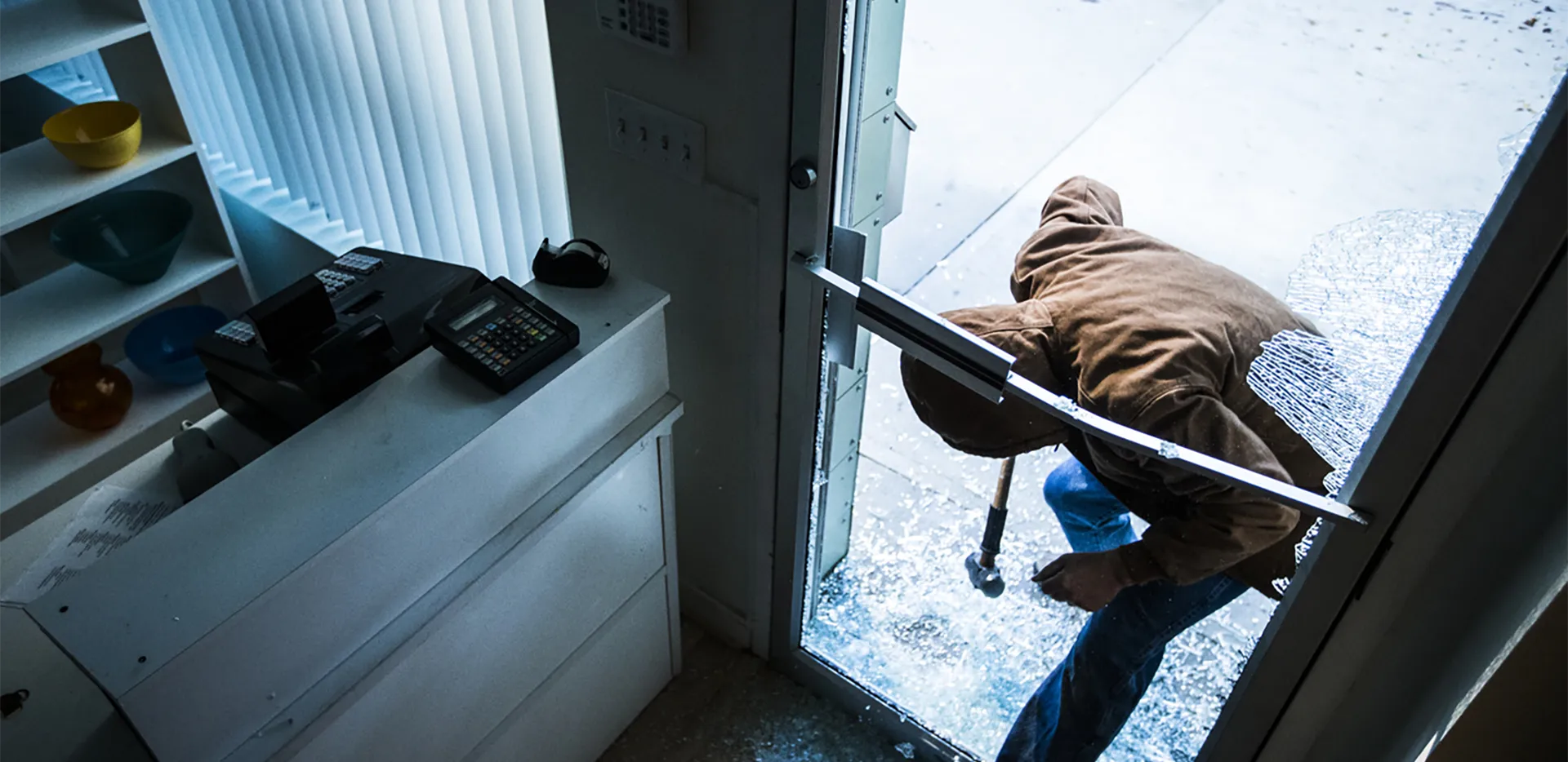 A person wearing a jacket breaking the front door of a business with a hammer.