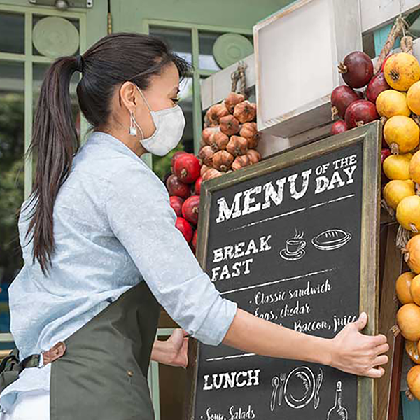 A business owner wearing a mask and placing a menu outside of their cafe.