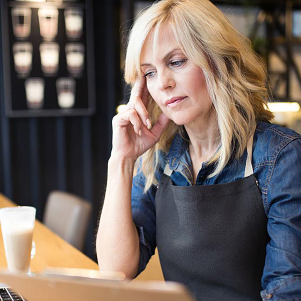A cafe owner wearing an apron clutching her head while working on her laptop.