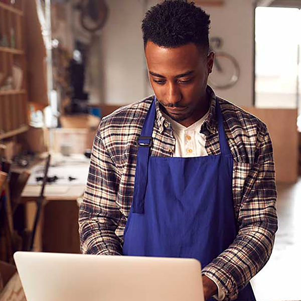 A man wearing an apron working on a laptop computer with construction equipment in the background.