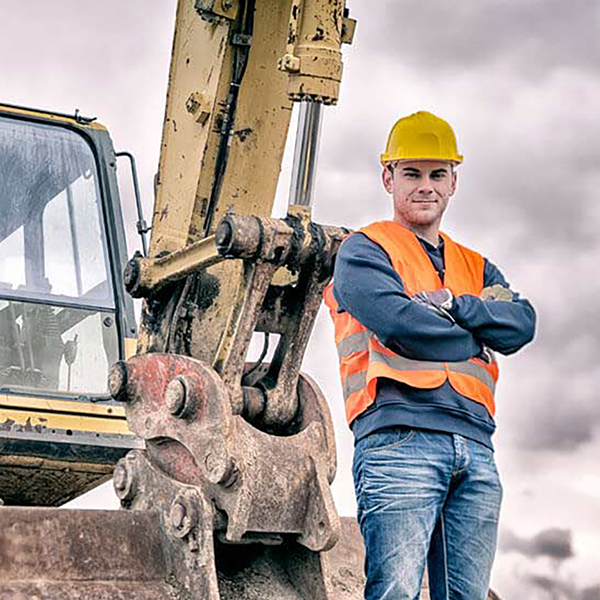 A construction worker standing with his arms folded in front of an excavator.