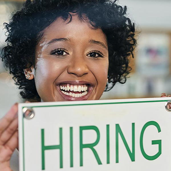 A smiling woman hanging a 'hiring' sign on the door of her business.