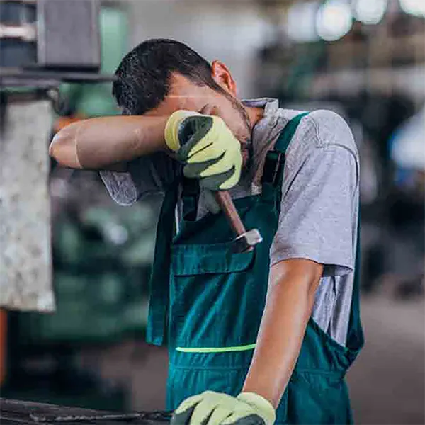A worker wearing an apron and gloves wiping sweat off of their forehead.