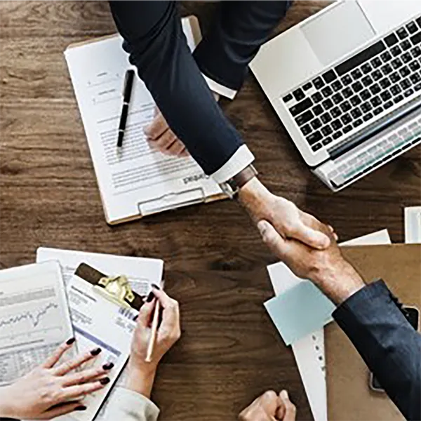 Two hands shaking against a background of documents and a laptop computer.