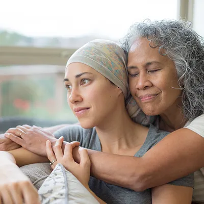 Two women embracing on a couch, one with a handkerchief tied over her head.