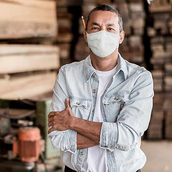 A man with a pencil over his ear wearing a mask. He's standing in front of a pile of lumber.