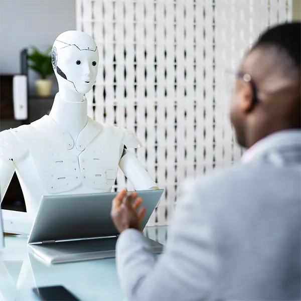 A robot sitting at a laptop speaks with a job candidate.