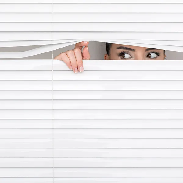 A woman peeking through the slats in a set of blinds.