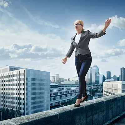 A woman balancing on the ledge of a tall building with a city behind her.