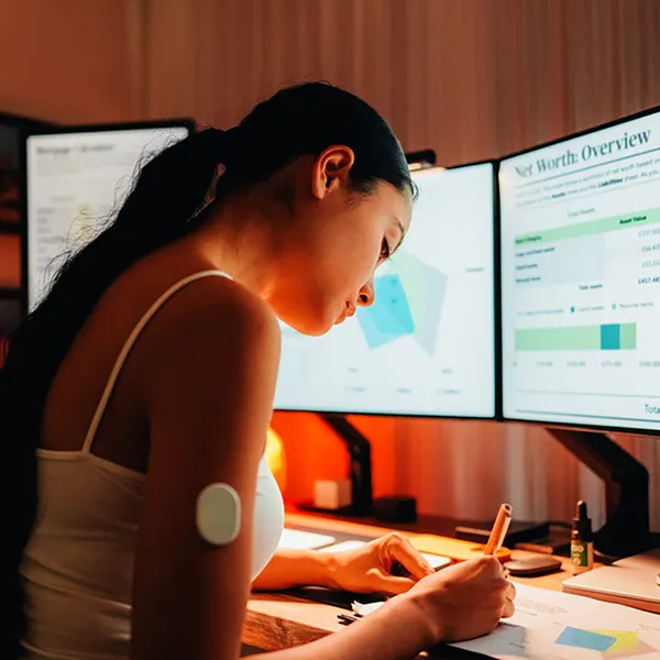 A woman taking notes while looking at graphs on a computer monitor.