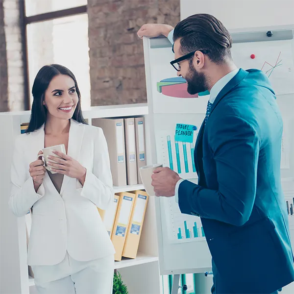 Two people dressed professionally smiling at each other while holding coffee cups.