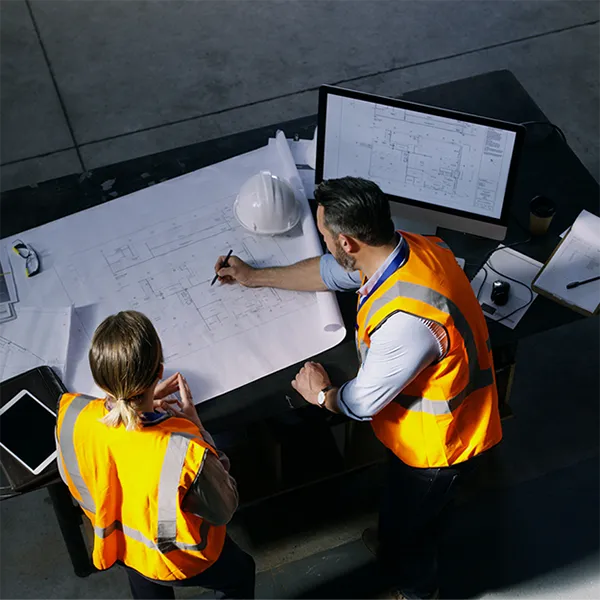 Two contractors in orange safety vests reviewing blueprints on a large desk.