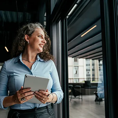 A smiling woman holding a tablet and looking across an office space.