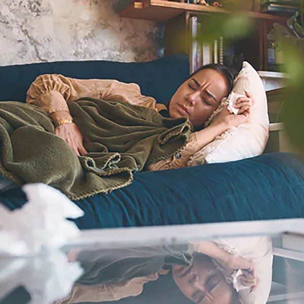 A person on their couch taking a nap. Tissues and a mug of tea can be seen in the foreground.