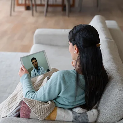 A woman in a blue sweater sitting on w white couch hold a tablet in her hand while on a telehealth call with a doctor.