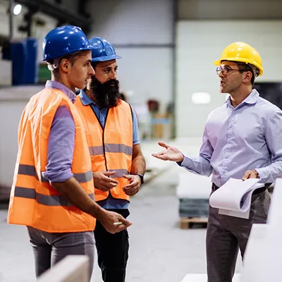 Three men in hard hats and safety gear having a discussion in an industrial space.