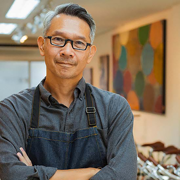 A bike shop owner wearing an apron looking at the camera with his arms crossed.