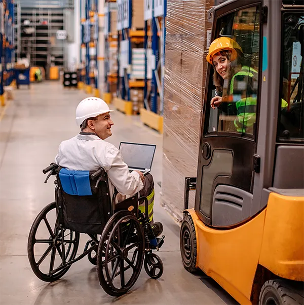 A worker using a wheelchair speaks with another worker driving a forklift. Both are wearing hard hats.