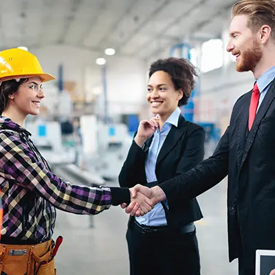 A man in a suit shaking hands with a woman wearing a hard had and tool belt.