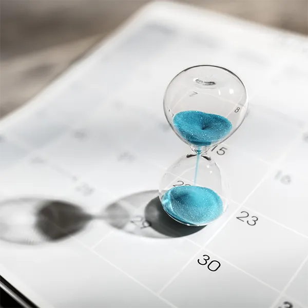 An hourglass with blue sand balanced on a calendar.