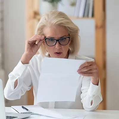 A woman holds her glasses and she reads a piece of paper in disbelief.