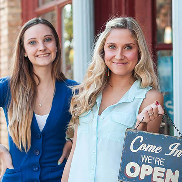 Two small business owners pose in front of their business holding a 'Come in we're open' sign.
