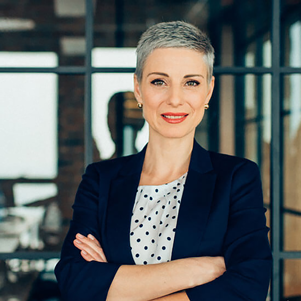 A professionally dressed woman standing with her arms folded in front of a glass window.