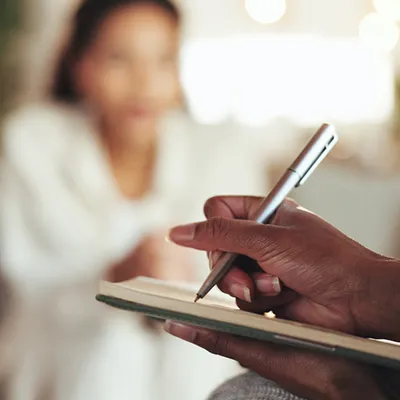 A hand writing in a notebook with the blurry shape of a woman in the background.